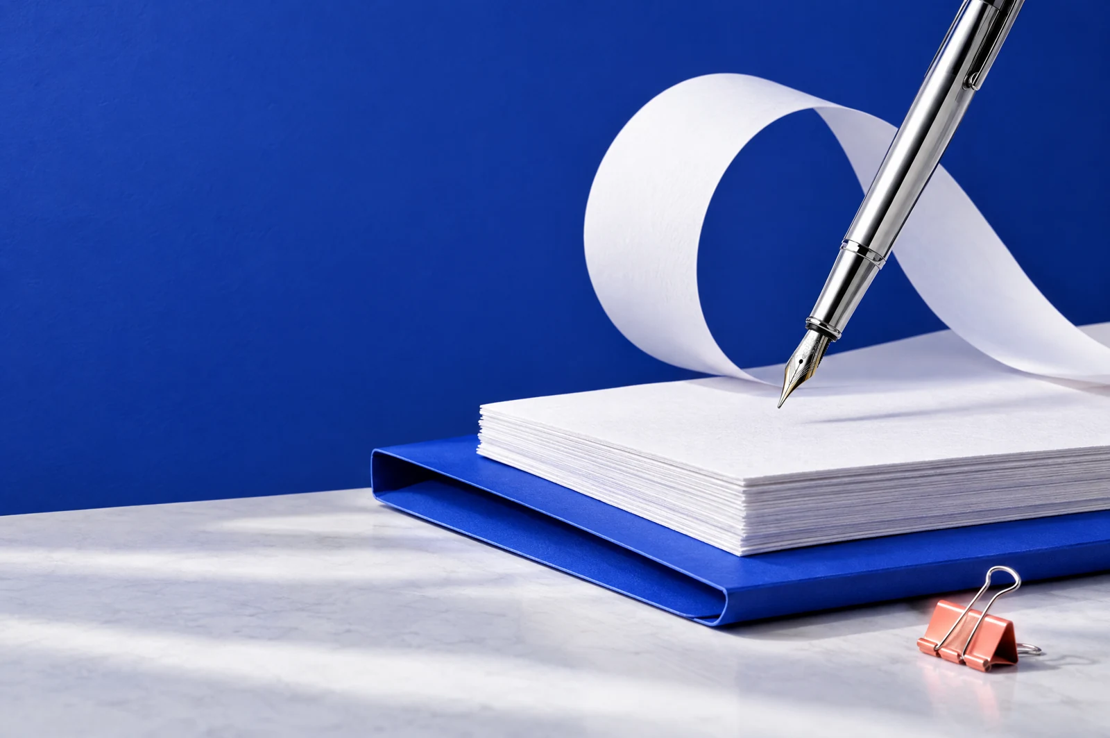 A silver fountain pen suspended above textured paper, a cobalt folder and a sculptural paper ribbon